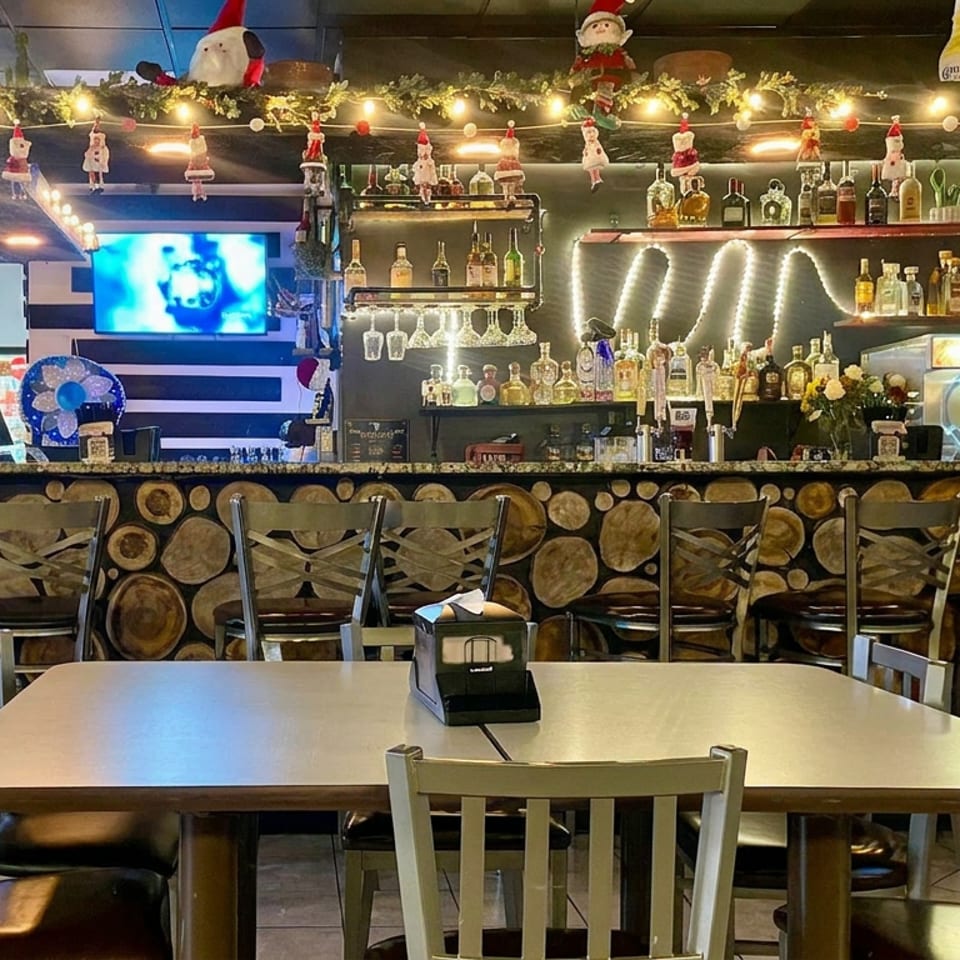 Festively decorated restaurant interior with a fully stocked bar, log-patterned counter, dining tables, televisions, and Christmas ornaments.