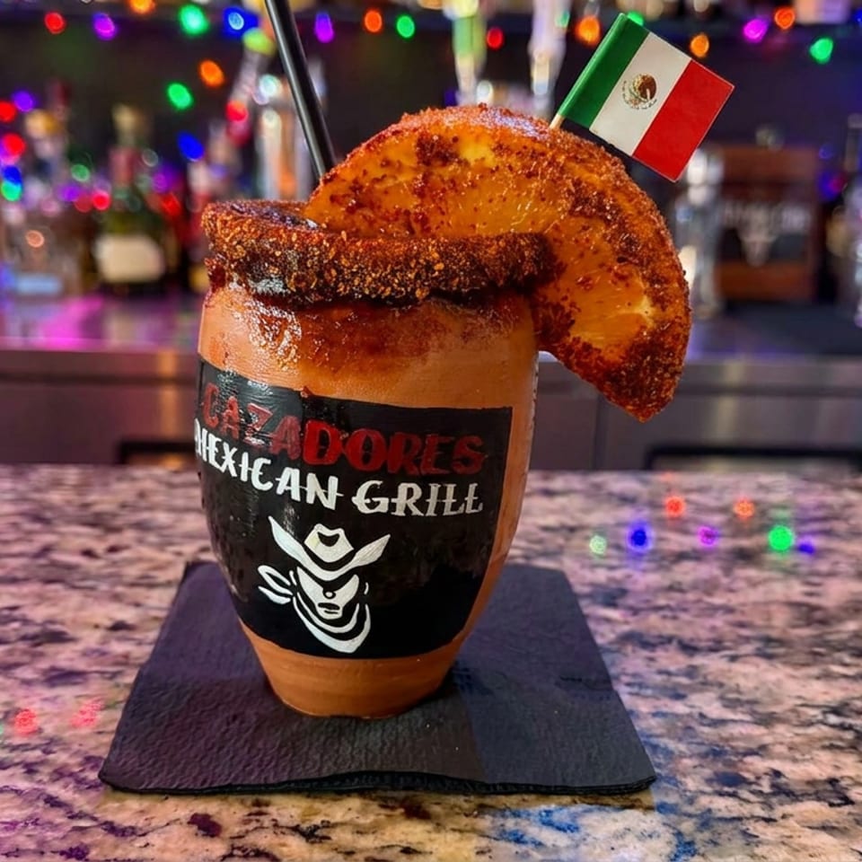 Mexican cocktail garnished with a chili-salt rim, dried citrus, and Mexican flag, displayed on a bar counter amid colorful lights.