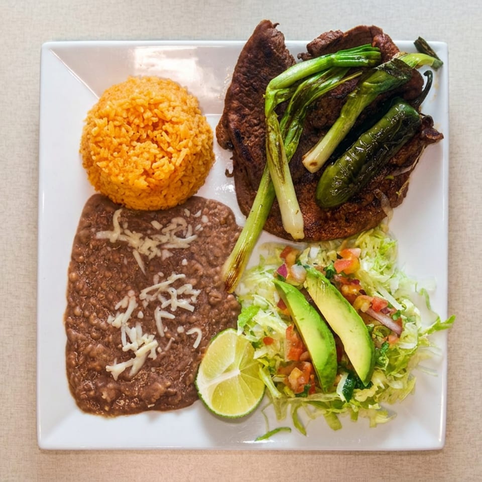 Mexican-style plate with grilled steak, sautéed vegetables, orange rice, refried beans, avocado salad, and lime wedges.