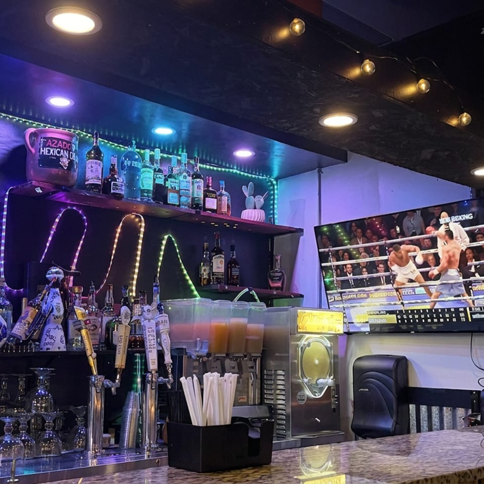 Colorful restaurant bar with liquor bottles, beer taps, drink dispensers, chalkboard specials, and a television showing wrestling.